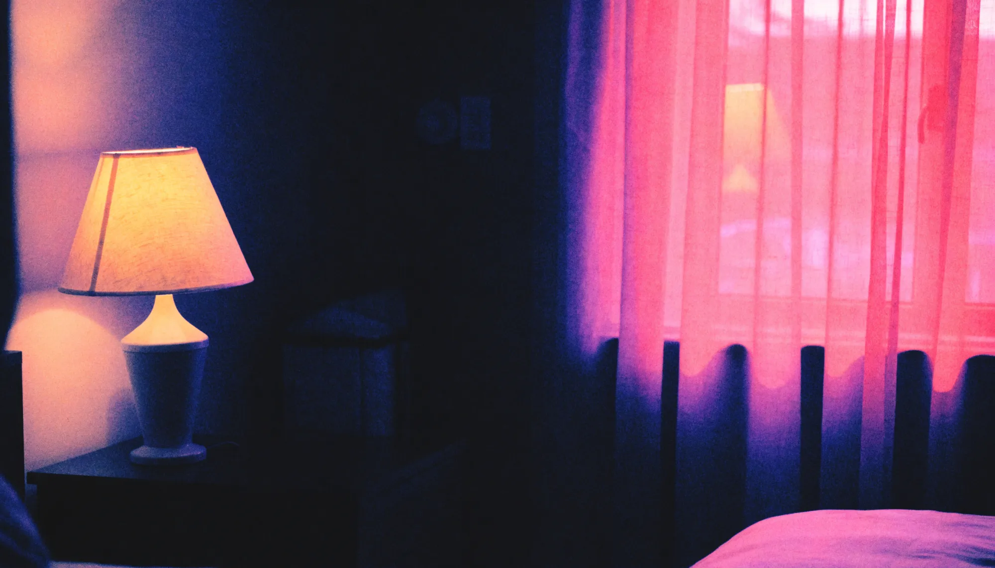 A dim bedroom lit by a warm lamp and soft pink light through sheer curtains at night