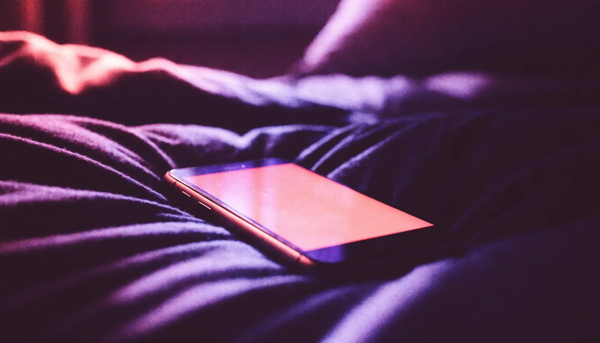 A phone glowing with soft pink screen light on dark bedsheets late at night